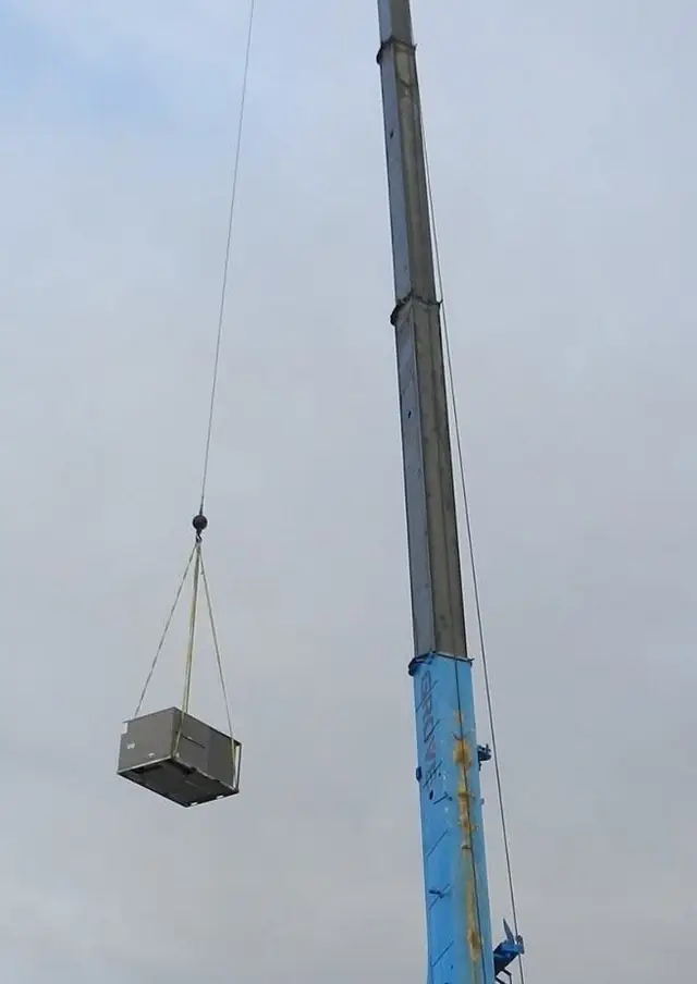A rooftop HVAC unit hanging from a crane boom against an overcast sky